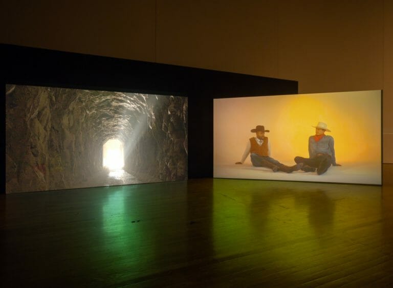 2 channel installation of Kenneth Tam: Silent Spikes with a tunnel on one screen and seated cowboys on the other