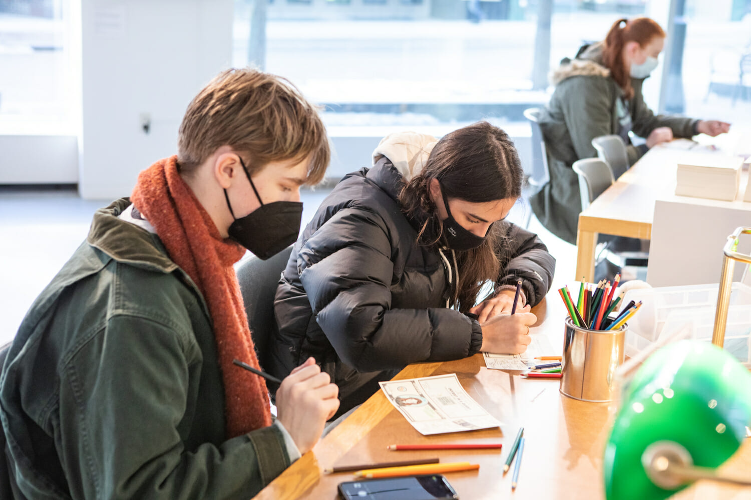 two teenagers or college students seated at a table, decorating Fundred Dollar Bills with color pencils