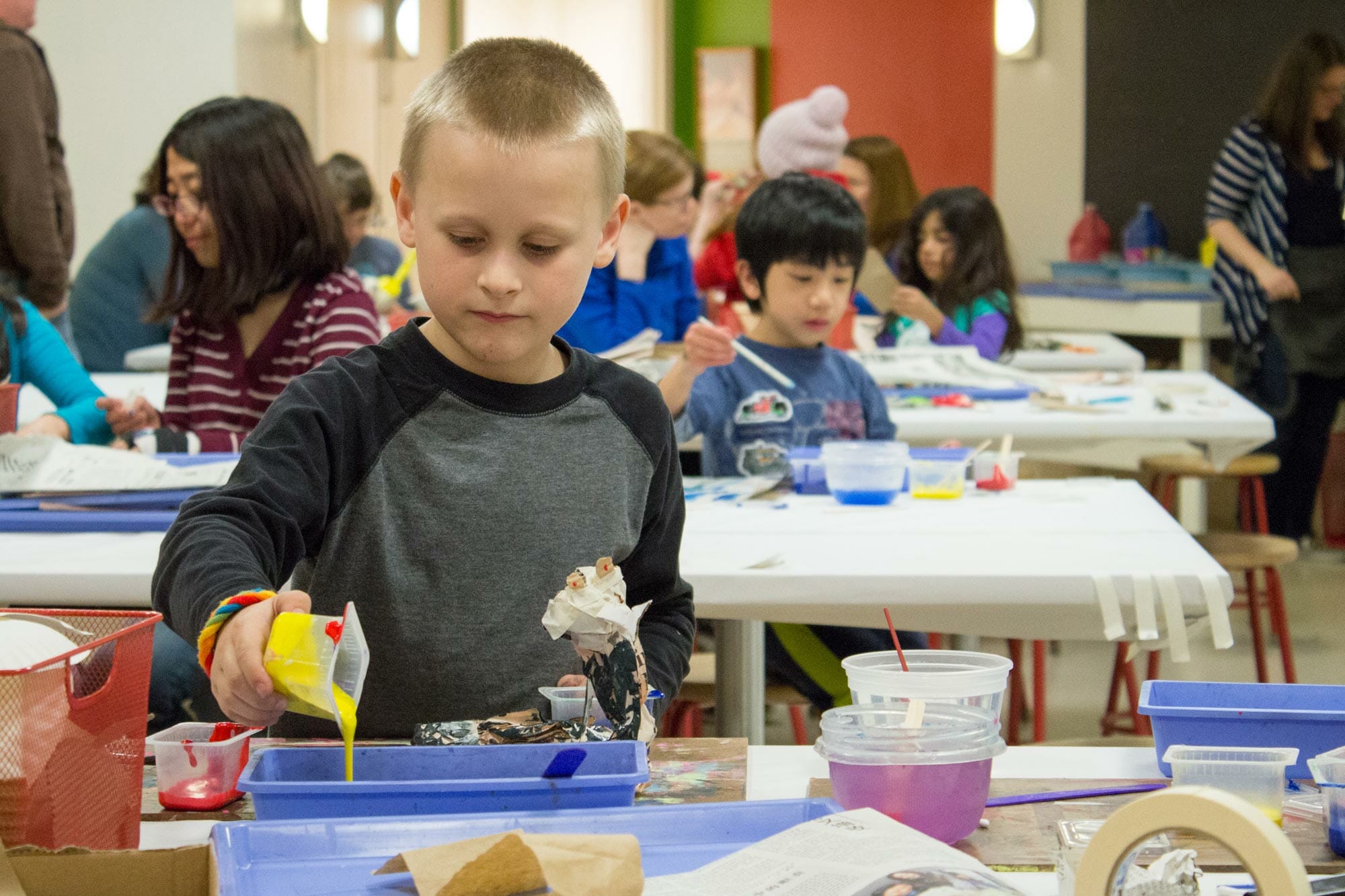 a child pouring paint from one container to a larger container. Behind this child are other children seated at rows of tables participating in an artistic activity