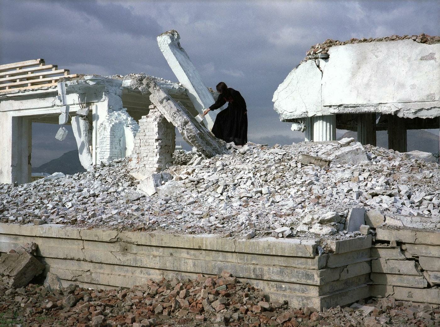 still from a video showing a person standing on a tall pile of rubble, wearing a dress, holding a wide paintbrush and brushing the surface of a collapsed concrete wall