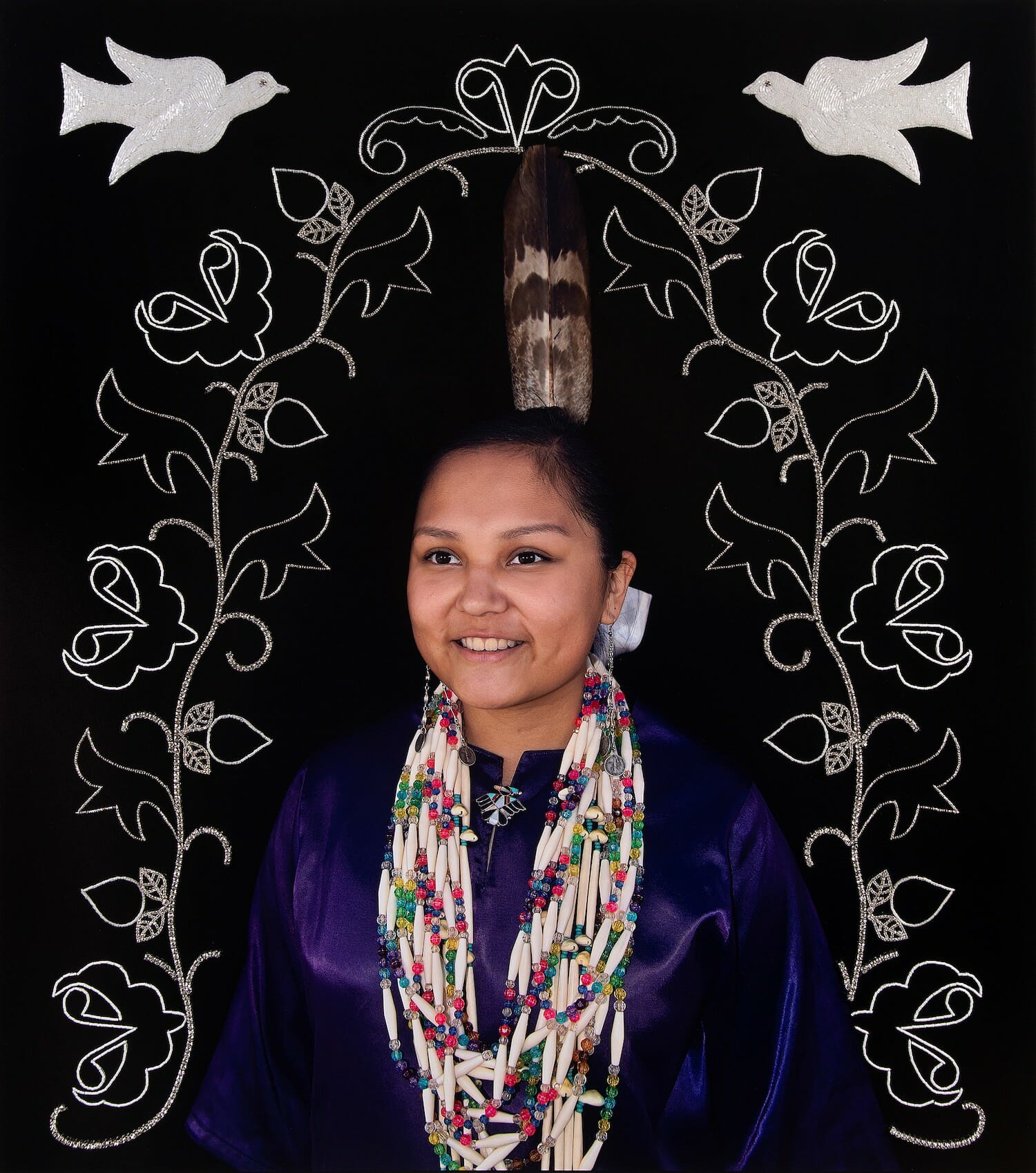 photograph of a Ho-Chunk girl in traditional clothing surrounded by floral beadwork and embroidery and embroidered two birds flying above her head