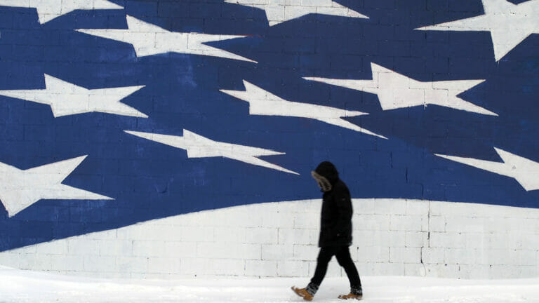 person walking, wearing a winder jacket, jeans, and boots walking along a wall with a mural of the star portion of the American flag