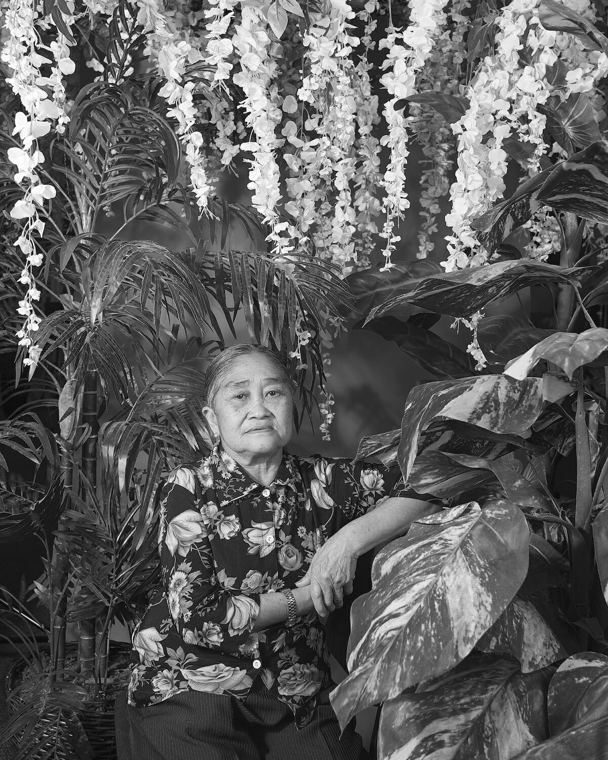 older person wearing a floral print button-up shirt, resting their arm against a surface, in a leafy, vegetal, floral setting