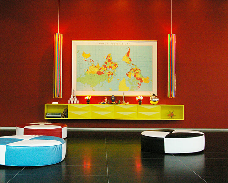 installation view of The Absent City, meant to be an interactive installation modeled as a public living room, with large checkerboard cushions, a floating credenza with various objects, and a poster of a world map