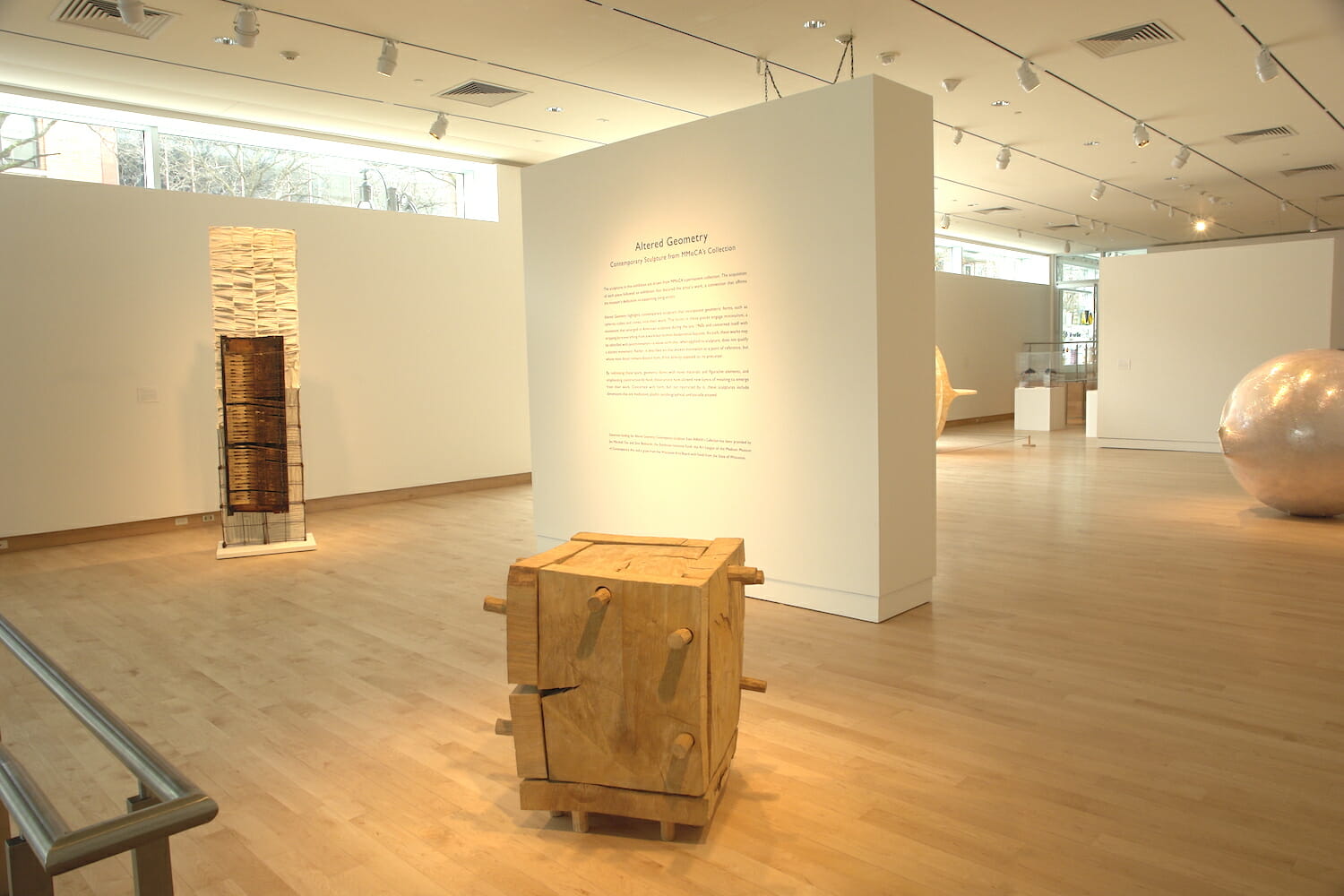 installation view of the exhibition Altered Geometry, featuring a stand alone wall with title wall text, surrounded by a wooden box sculpture, a tall abstract sculpture, a round sculpture with a plastic exterior