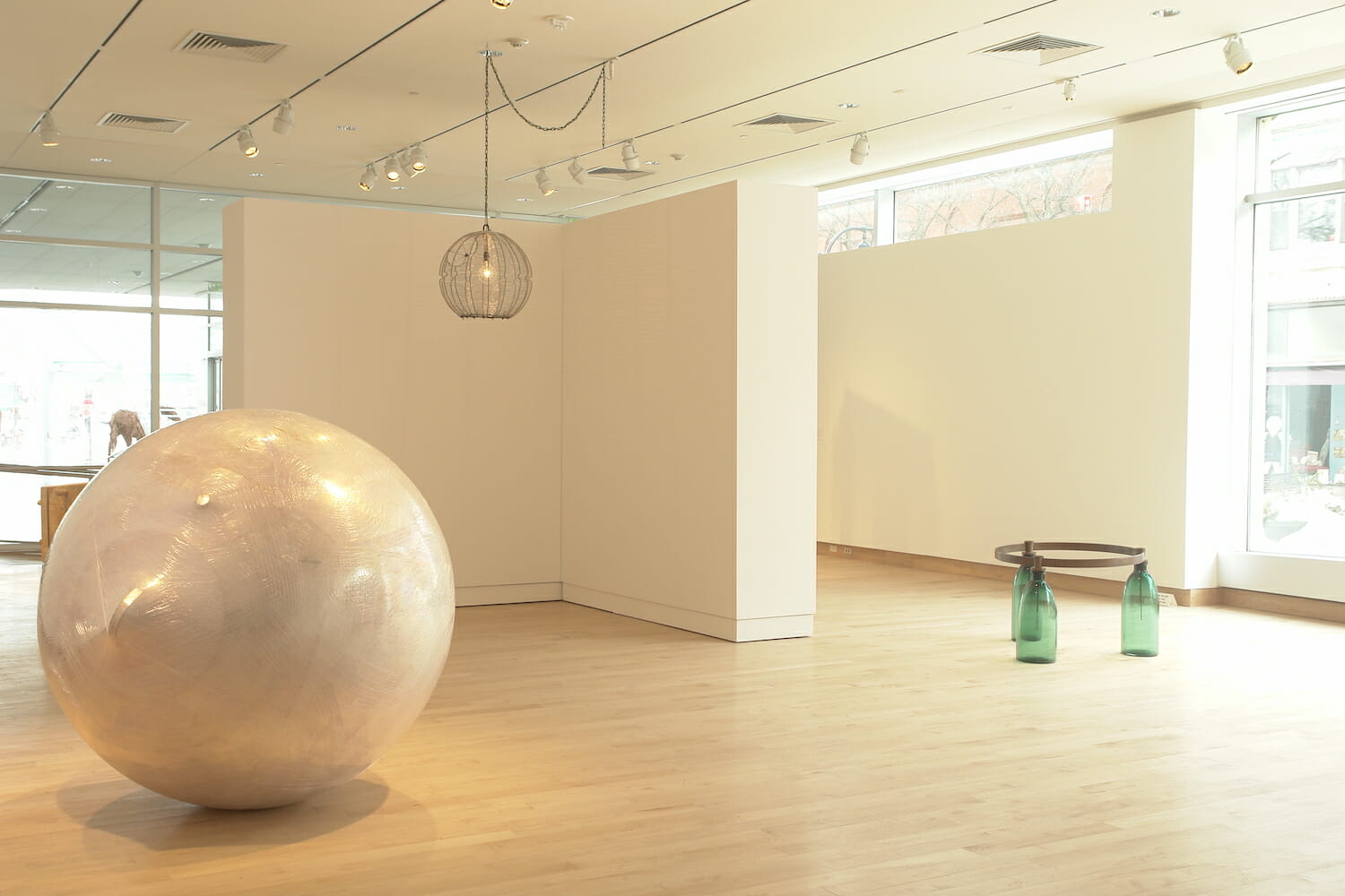 installation view of the exhibition Altered Geometry, featuring a round abstract sculpture, a hanging light fixture, and another abstract sculpture with a round metal ring balanced on top of three green bottles