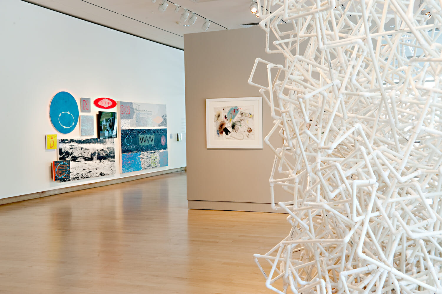 gallery installation view of the 2010 Wisconsin Triennial showing a partial close up of a sculpture to the front right and additional rectangular artworks hanging on a wall