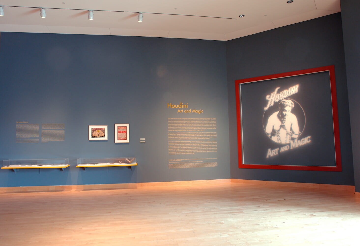 installation view of Houdini: Art and Magic featuring framed artworks, title wall text, and floating vitrine displays with various artworks or objects