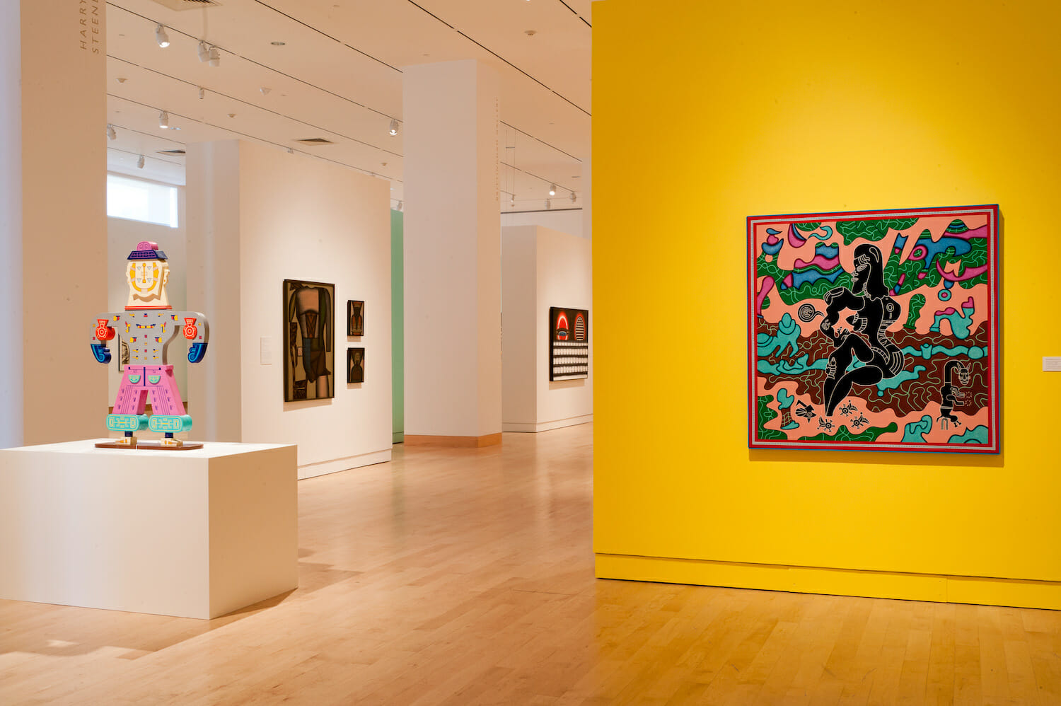 installation view of the exhibition Chicago Imagists featuring a painted wooden cutout shaped like a paper doll on a display platform. painted canvases or other artwork are hung on nearby walls.