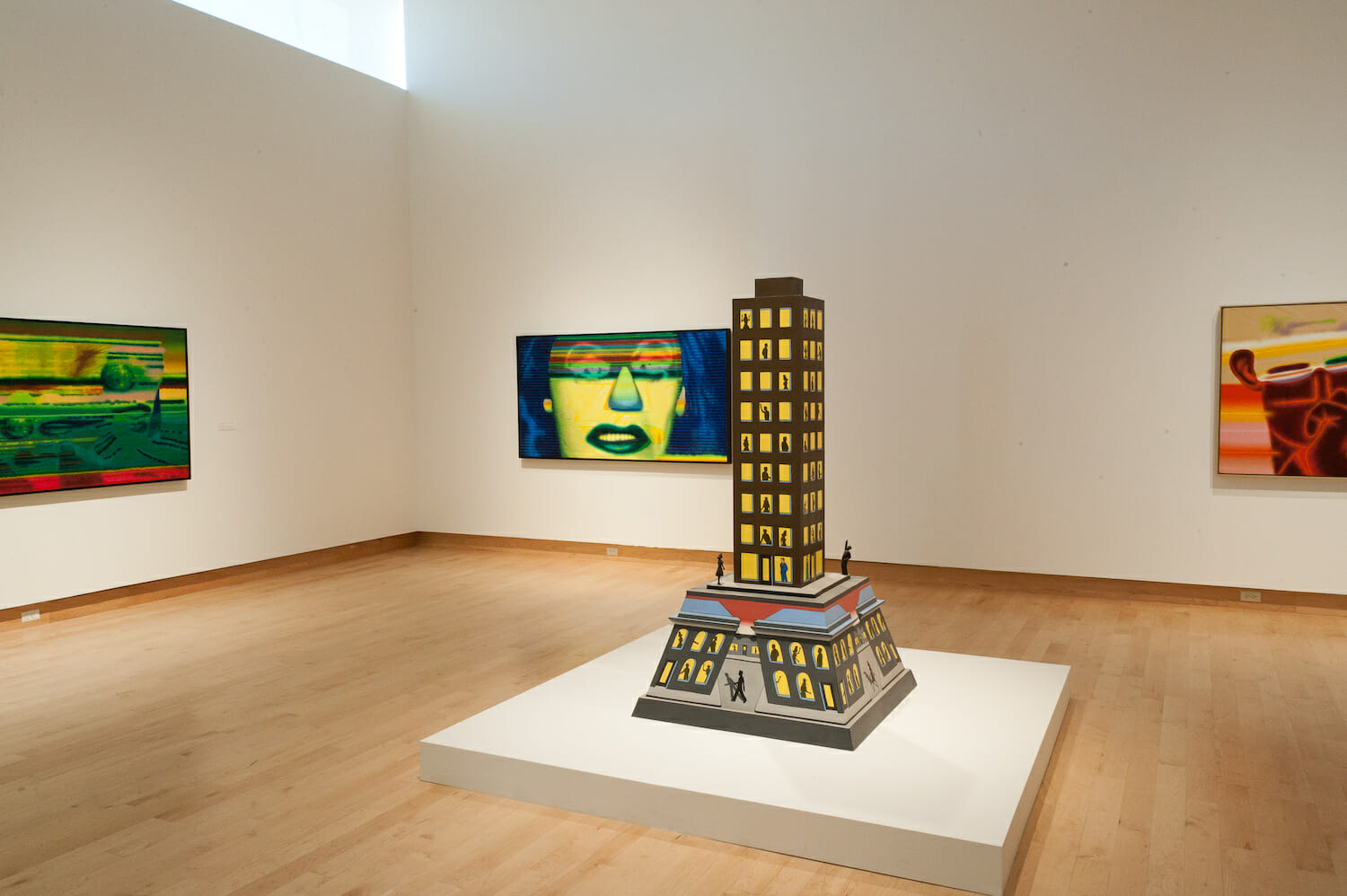 installation view of the exhibition Chicago Imagists featuring a painted wooden sculpture of a building, painted in a cartoonish style. three painted canvases or artworks are displayed on the walls surrounding the sculpture.