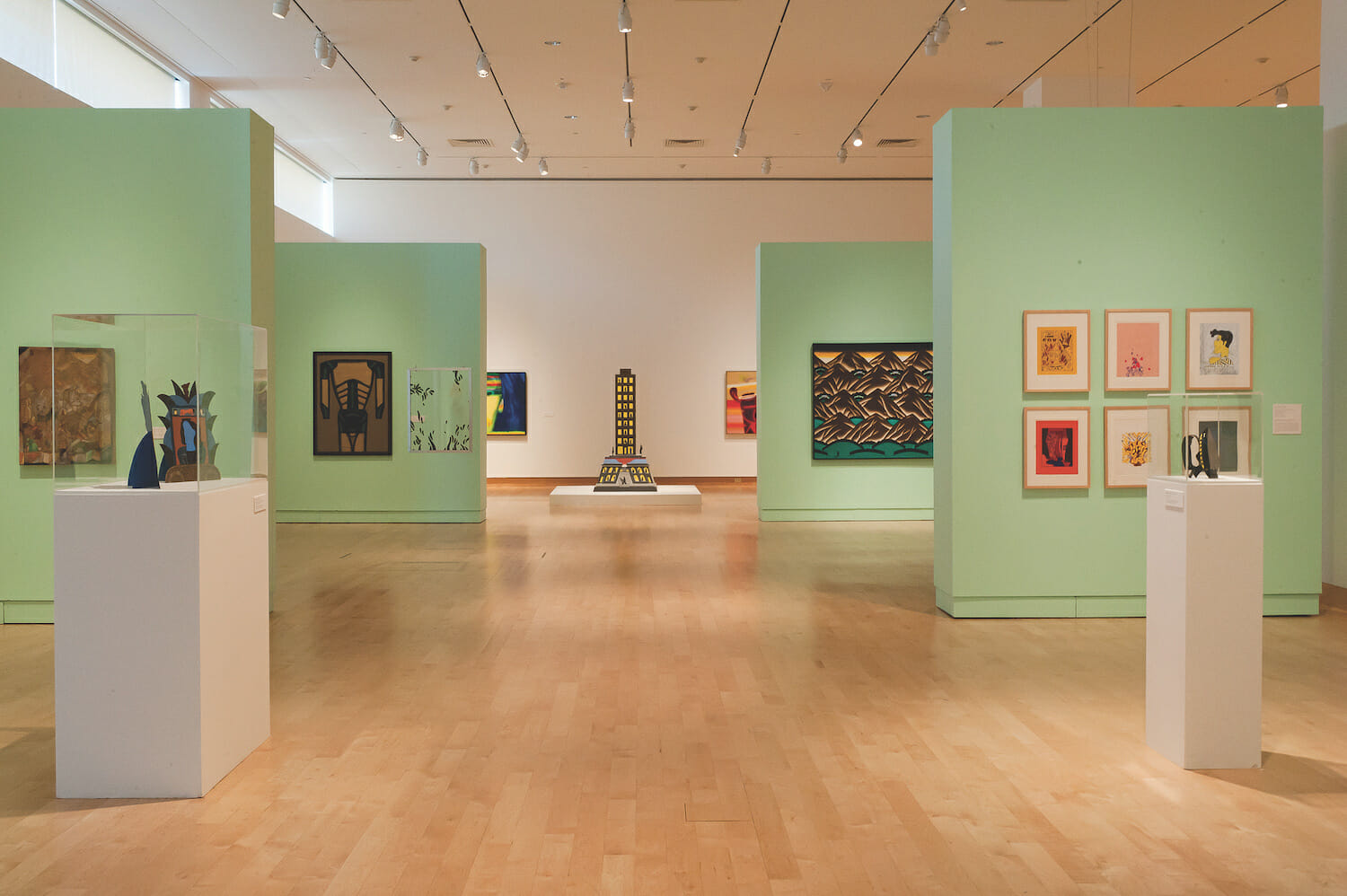 gallery installation view of the exhibition Chicago Imagists. two vitrine display cases contain various artworks or objects. artworks hang on several different walls. a tall painted sculpture resembling a building stands on a platform towards the back center of the room.