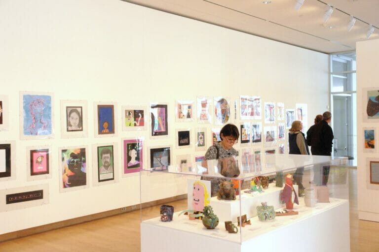 installation view of the 2013 Young at Art exhibition featuring rows of artworks by children on display across a wall and a vitrine display case featuring smaller sculptures or ceramics