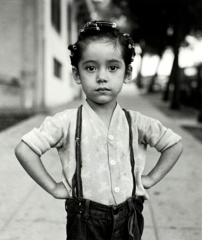 child with curlers in her hair and her fists on her hips, wearing a button-up shirt and pants with suspenders