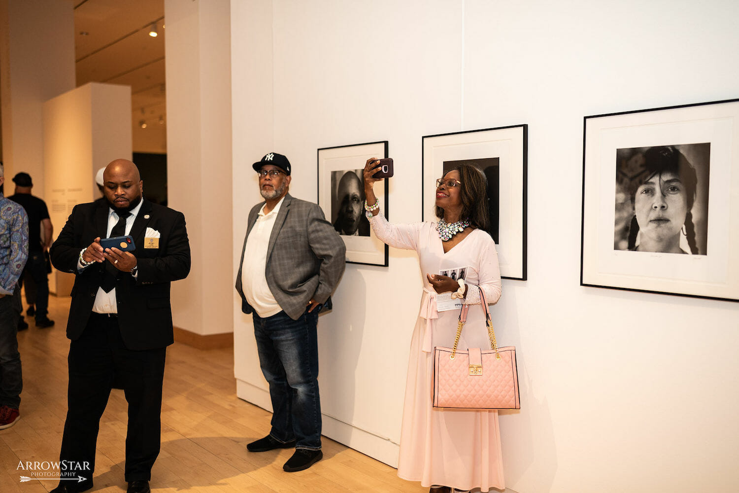 A person holds their phone up to take a photo. Behind them are framed photographs on display across a wall. Another person looks at their phone, possibly at a photo they had just taken. A person nearby stands, about to tuck their hands in their pocket.