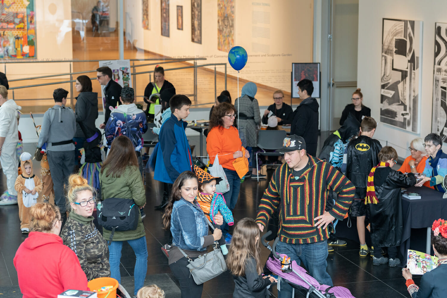 A crowd of people in the Museum Lobby, consisting of family and children dressed up in Halloween costumes, enjoying a variety of treats and activities.