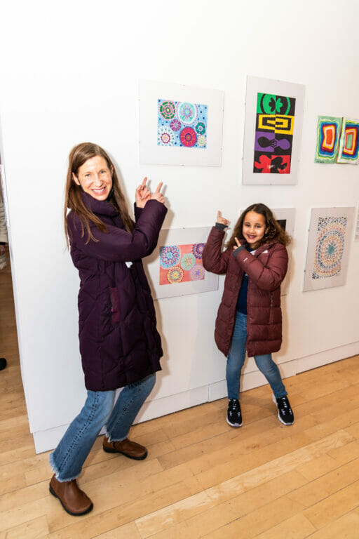 An adult and a student artist poses for a photo while pointing at the student's artwork which is hanging on a wall on display.