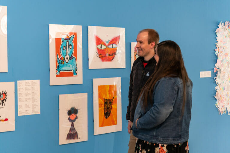 Two adults look at children's artwork which are hanging across a wall in an exhibition.