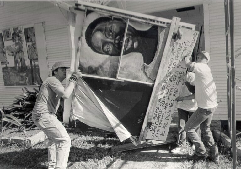 Three people moving a giant painted artwork in front of a house outdoors.