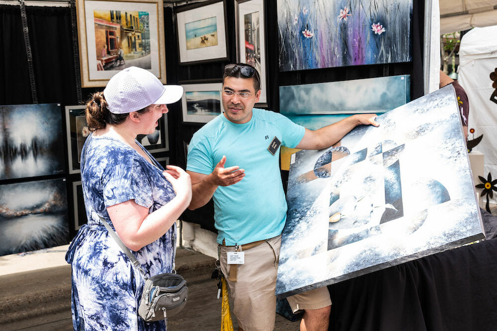 Artist holding a large art canvas talking to an art fair visitor.