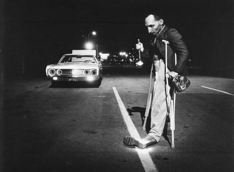 Black and white image of a figure in the road with crutches and an automobile behind them.