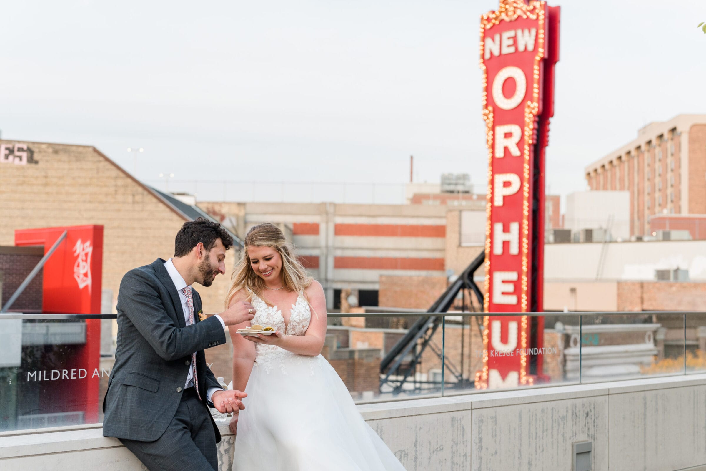 A man in a suit and a woman in a wedding dress eating cake with the New Orpheum sign behind them.