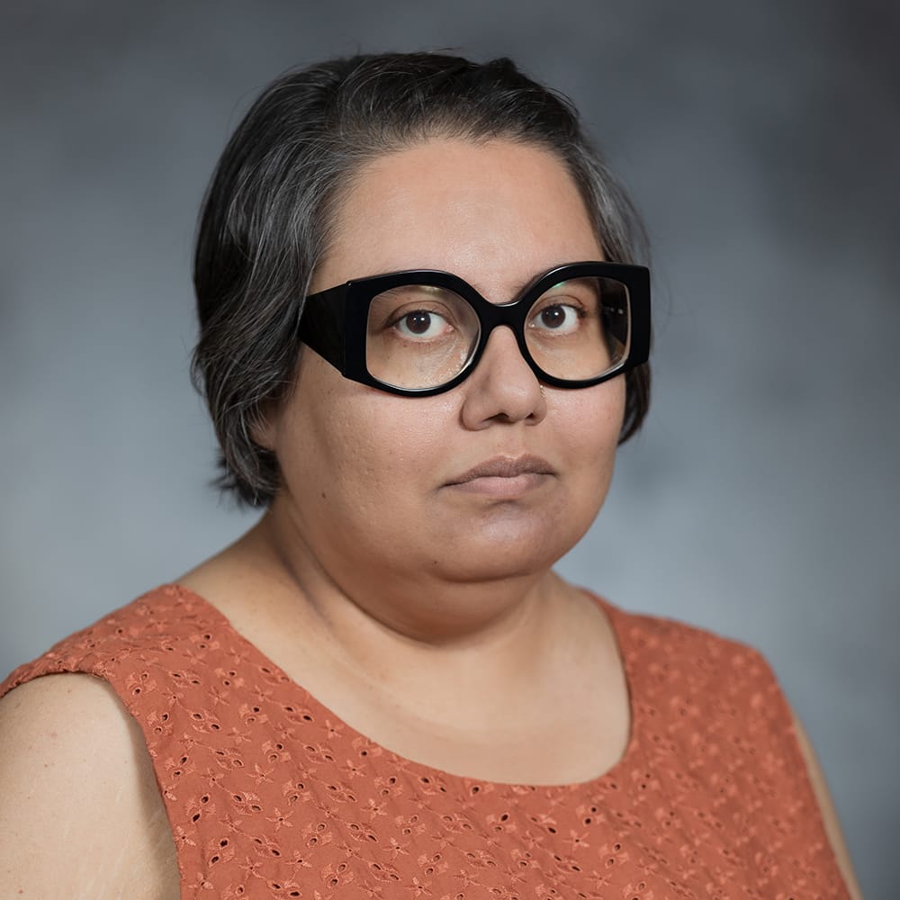 Portrait of Dr. Mou Banerjee wearing thick black glasses and an orange shirt against a gray background.