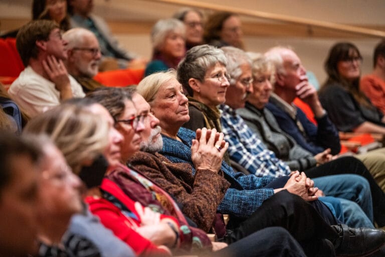 Multiple people inside of a lecture hall with red chairs.