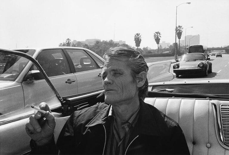 A black and white film still of Chet Baker sitting in the back of a convertible on a highway with the top down smoking a cigarette with his eyes closed.