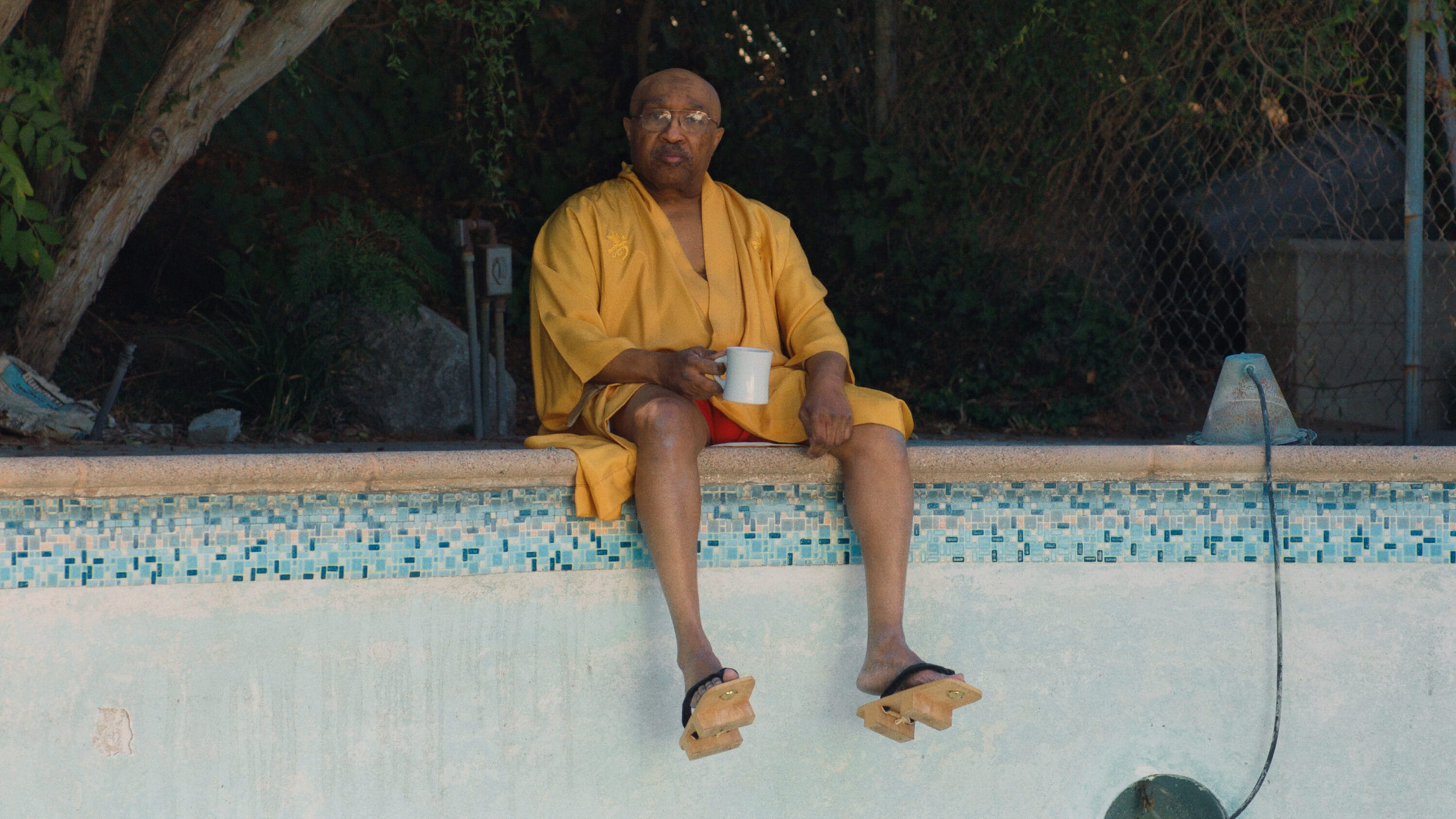 A black man sits on the edge of an empty swimming pool holding a mug and wearing a yellow robe.