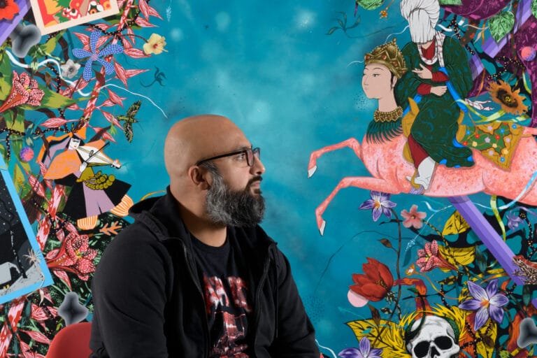 A portrait of Amir H. Fallah. A man with a black beard, shaved head, and glasses wearing a black sweatshirt in front of a painting with a blue backgroun.